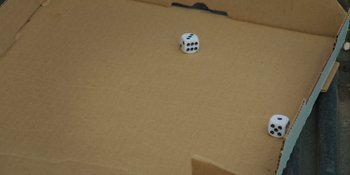 Movie still from “The Retirement Plan” (2023), directed by Tim Brown – A dice sitting on top of a cardboard box on the floor; Extreme Close Up shot, High angle