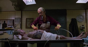 Movie still from “The Return of the Living Dead” (1985), directed by Dan O'Bannon – A man is performing surgery on an older man; Medium shot, Over the shoulder angle