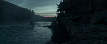 Movie still from “The Revenant” (2015), directed by Alejandro G. Iñárritu – A man sitting on a rock near a body of water; Extreme Wide shot, High angle