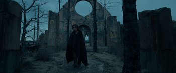 Movie still from “The Revenant” (2015), directed by Alejandro G. Iñárritu – A man in a black cloak standing in front of a building; Wide shot, Low angle