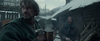 Movie still from “The Revenant” (2015), directed by Alejandro G. Iñárritu – Two men sitting in front of a hut drinking from a cup; Medium shot, Low angle
