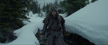 Movie still from “The Revenant” (2015), directed by Alejandro G. Iñárritu – A man with a beard and a fur hat is walking through the snow; Wide shot, Low angle
