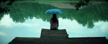 Movie still from “The Ritual Killer” (2023), directed by George Gallo – A woman sitting on a dock holding an umbrella; Wide shot, High angle