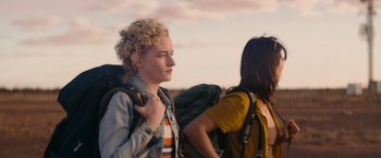 Movie still from “The Royal Hotel” (2023), directed by Kitty Green – Two people with backpacks are standing outside; Medium shot, Over the shoulder angle