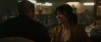 Movie still from “The Royal Hotel” (2023), directed by Kitty Green – A woman smiling at the camera while sitting next to a man; Close Up shot, Over the shoulder angle