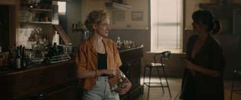 Movie still from “The Royal Hotel” (2023), directed by Kitty Green – A woman standing in front of a bar holding a bottle of beer; Medium shot, Over the shoulder angle