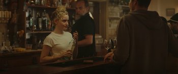 Movie still from “The Royal Hotel” (2023), directed by Kitty Green – A woman sitting at a bar holding a bottle of wine; Medium shot, Over the shoulder angle