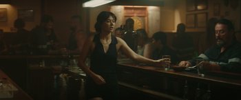 Movie still from “The Royal Hotel” (2023), directed by Kitty Green – A woman standing in front of a bar holding a glass; Medium shot, Over the shoulder angle