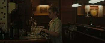 Movie still from “The Royal Hotel” (2023), directed by Kitty Green – A woman is standing at a counter with glasses on it; Medium shot, Over the shoulder angle