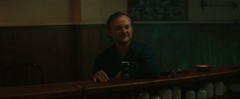 Movie still from “The Royal Hotel” (2023), directed by Kitty Green – A man sitting at a table with a can of beer; Medium shot, Over the shoulder angle