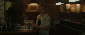 Movie still from “The Royal Hotel” (2023), directed by Kitty Green – A woman standing in front of a counter with wine glasses on it; Medium shot, Over the shoulder angle