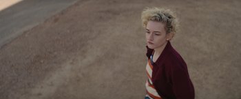 Movie still from “The Royal Hotel” (2023), directed by Kitty Green – A woman with blonde curly hair standing on a dirt field; Close Up shot, High angle