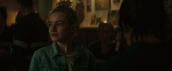 Movie still from “The Royal Hotel” (2023), directed by Kitty Green – A woman with a messy bun is standing in front of a group of people; Close Up shot, Over the shoulder angle