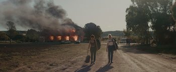 Movie still from “The Royal Hotel” (2023), directed by Kitty Green – Two women walking down a dirt road near a burning building; Extreme Wide shot, Low angle