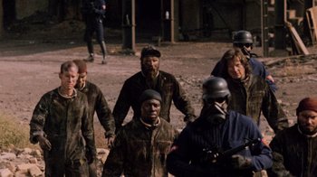 Movie still from “The Running Man” (1987), directed by Paul Michael Glaser – A group of men in a field with a person in a gas mask; Medium shot, High angle