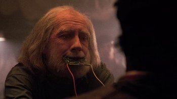 Movie still from “The Running Man” (1987), directed by Paul Michael Glaser – An old man with a beard and wires in his mouth; Close Up shot, Over the shoulder angle