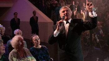 Movie still from “The Running Man” (1987), directed by Paul Michael Glaser – A man in a black suit is speaking into a microphone; Medium shot, Over the shoulder angle