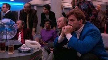 Movie still from “The Running Man” (1987), directed by Paul Michael Glaser – A group of people sitting around a table; Medium shot, Over the shoulder angle
