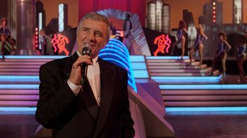 Movie still from “The Running Man” (1987), directed by Paul Michael Glaser – An older man in a black suit is holding a microphone; Medium shot, Low angle