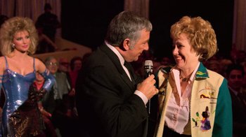 Movie still from “The Running Man” (1987), directed by Paul Michael Glaser – An older man and a woman sing into microphones at an event; Medium shot, Over the shoulder angle