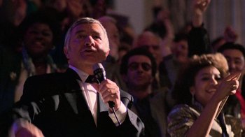 Movie still from “The Running Man” (1987), directed by Paul Michael Glaser – An older man is holding a microphone in front of an audience; Medium shot, Over the shoulder angle