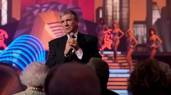 Movie still from “The Running Man” (1987), directed by Paul Michael Glaser – A man in a black suit is holding a microphone; Medium shot, Over the shoulder angle