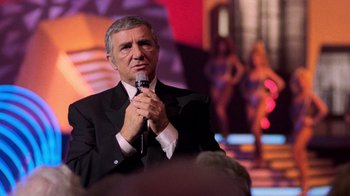 Movie still from “The Running Man” (1987), directed by Paul Michael Glaser – A man in a suit and tie holding a microphone; Medium shot, Over the shoulder angle