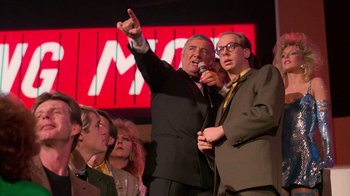 Movie still from “The Running Man” (1987), directed by Paul Michael Glaser – A group of people standing around a microphone on a stage; Medium shot, Over the shoulder angle