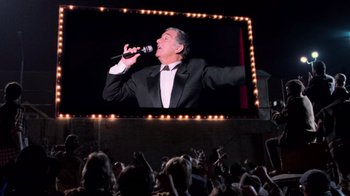 Movie still from “The Running Man” (1987), directed by Paul Michael Glaser – A man in a suit is singing in front of a large screen; Medium shot, Low angle