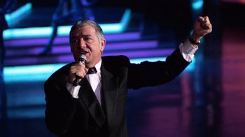 Movie still from “The Running Man” (1987), directed by Paul Michael Glaser – A man in a black suit is singing into a microphone; Medium shot, Low angle