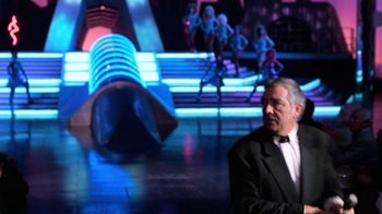 Movie still from “The Running Man” (1987), directed by Paul Michael Glaser – A man in a suit and tie in front of neon lights; Wide shot, Over the shoulder angle