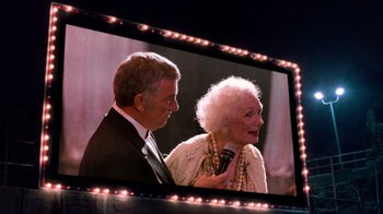 Movie still from “The Running Man” (1987), directed by Paul Michael Glaser – An older man standing next to an older woman in front of a television screen; Medium shot, High angle