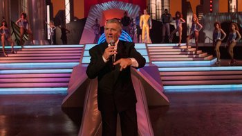 Movie still from “The Running Man” (1987), directed by Paul Michael Glaser – A man in a black suit is holding a microphone; Wide shot, Over the shoulder angle