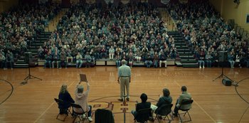 Movie still from “Dopesick” (2021), directed by Danny Strong – A man standing in front of an audience in a gymnasium; Extreme Wide shot, High angle