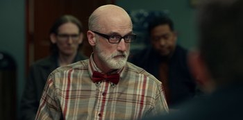 Movie still from “Dopesick” (2021), directed by Danny Strong – An older man wearing a bow tie in front of a group of people; Medium shot, Over the shoulder angle