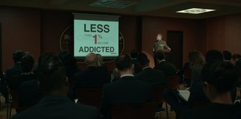 Movie still from “Dopesick” (2021), directed by Danny Strong – A man standing in front of a crowd of people in front of a projector screen; Extreme Wide shot, High angle