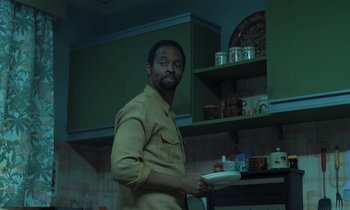 Movie still from “The Silent Twins” (2022), directed by Agnieszka Smoczynska – A man standing in a kitchen holding a white plate; Medium shot, Low angle