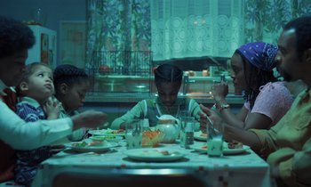 Movie still from “The Silent Twins” (2022), directed by Agnieszka Smoczynska – A group of people sitting at a table eating food; Medium shot, Low angle