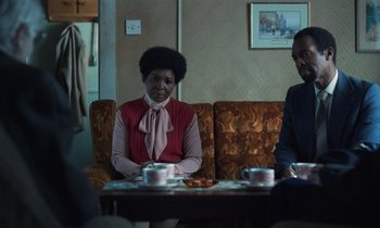 Movie still from “The Silent Twins” (2022), directed by Agnieszka Smoczynska – Two people sitting at a table in a living room; Medium shot, Over the shoulder angle