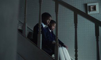 Movie still from “The Silent Twins” (2022), directed by Agnieszka Smoczynska – Two young people sitting on the stairs of a building; Wide shot, High angle