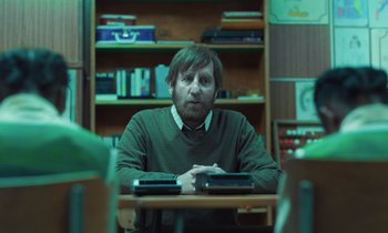 Movie still from “The Silent Twins” (2022), directed by Agnieszka Smoczynska – A man sitting at a table in front of a book shelf; Medium shot, Over the shoulder angle