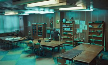 Movie still from “The Silent Twins” (2022), directed by Agnieszka Smoczynska – A man standing at a table in a room with two children; Wide shot, High angle