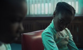 Movie still from “The Silent Twins” (2022), directed by Agnieszka Smoczynska – A young boy sitting on a chair in a room; Close Up shot, Over the shoulder angle