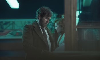 Movie still from “The Silent Twins” (2022), directed by Agnieszka Smoczynska – A man wearing a suit and tie looking down; Medium shot, Over the shoulder angle