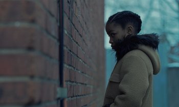 Movie still from “The Silent Twins” (2022), directed by Agnieszka Smoczynska – A young girl wearing a coat leaning against a brick wall; Close Up shot, Over the shoulder angle