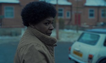 Movie still from “The Silent Twins” (2022), directed by Agnieszka Smoczynska – A woman in a brown jacket standing next to a white car; Close Up shot, Over the shoulder angle