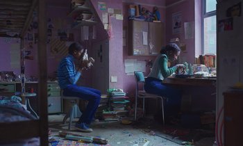 Movie still from “The Silent Twins” (2022), directed by Agnieszka Smoczynska – A couple of people sitting at a table in a room; Wide shot, Low angle