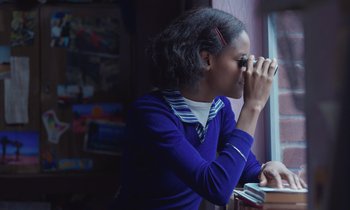 Movie still from “The Silent Twins” (2022), directed by Agnieszka Smoczynska – A woman looking out of a window at a window; Close Up shot, Over the shoulder angle