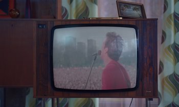 Movie still from “The Silent Twins” (2022), directed by Agnieszka Smoczynska – A man with a microphone on a television screen; Medium shot, High angle