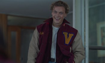 Movie still from “The Silent Twins” (2022), directed by Agnieszka Smoczynska – A young man in a varsity jacket smiling for the camera; Medium shot, Low angle